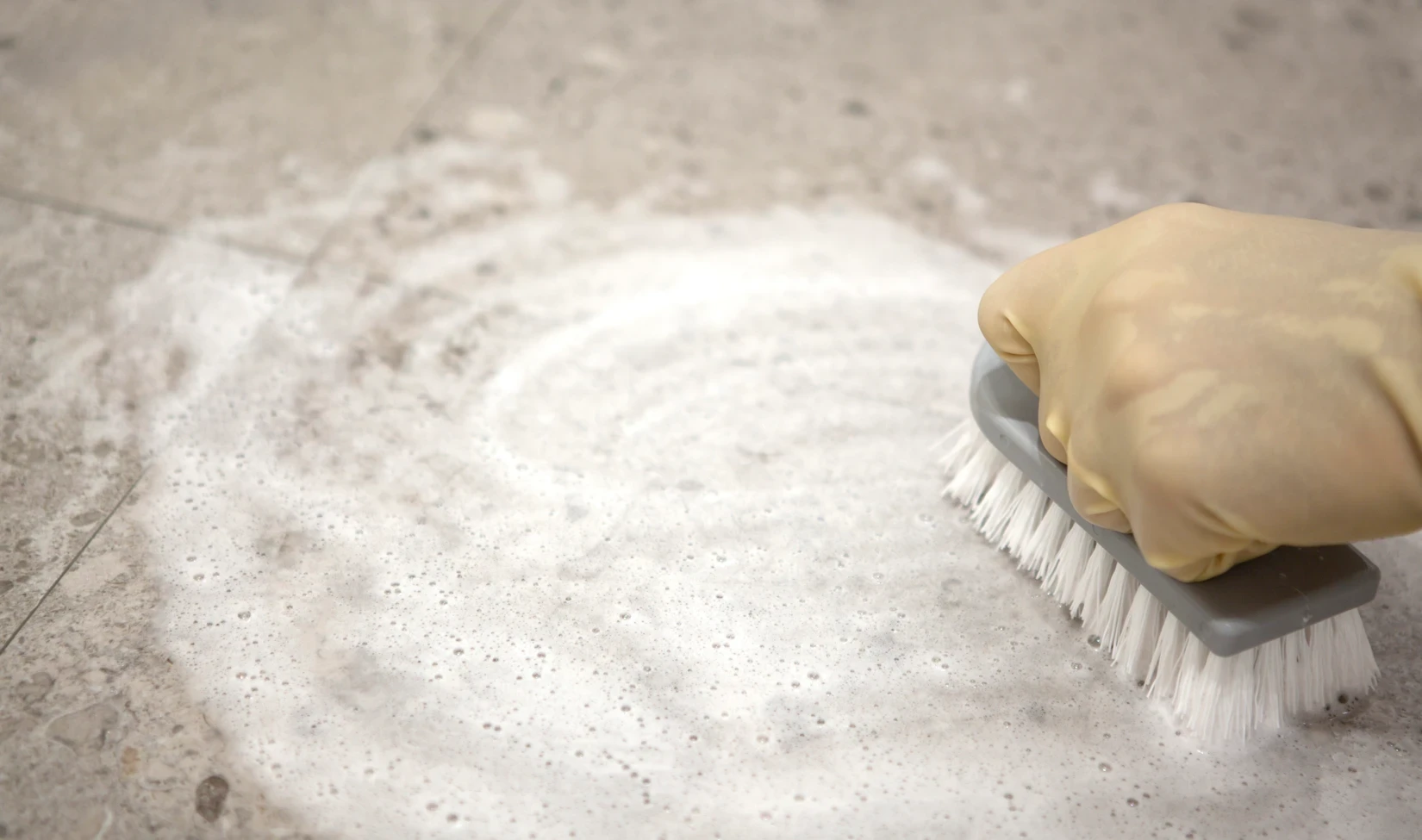 Main dans un gant protecteur jaune nettoyant le sol en grès cérame avec un détergent moussant et une brosse, concept d'entretien du sol carrelé.